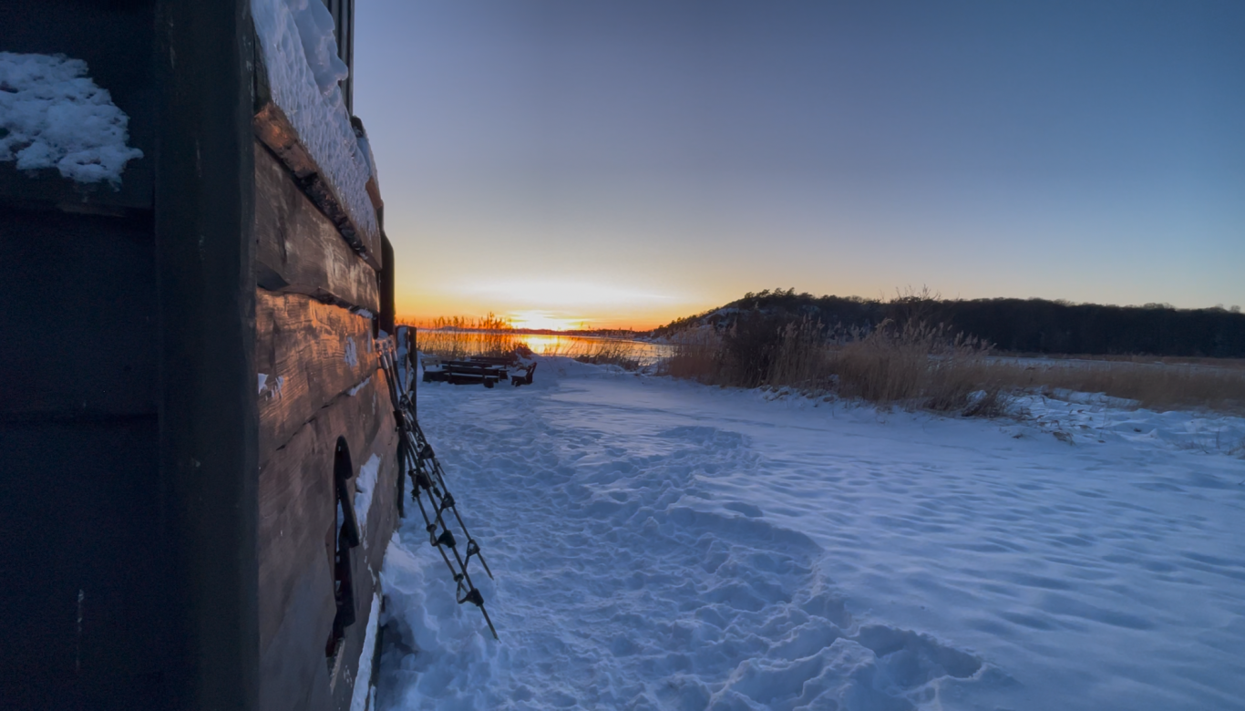 Skeppet Monterose Tjolöholms Slott Vinterlandskap vid vatten med snö, trästruktur i förgrunden och solnedgång i bakgrunden.
