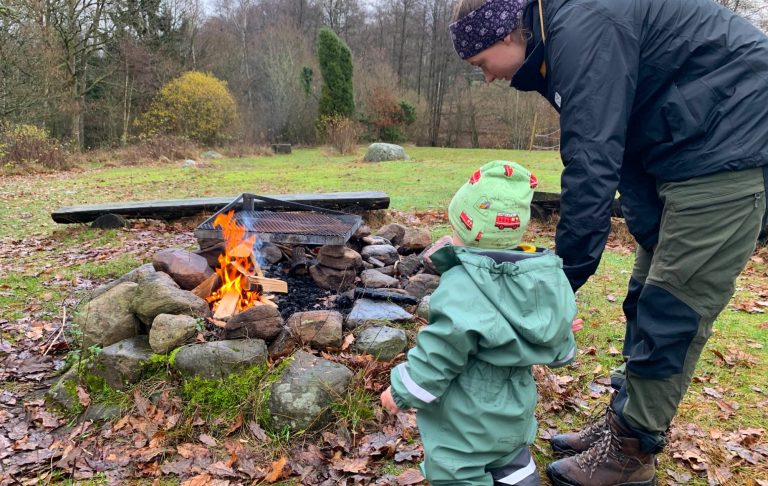 En vuxen och ett barn står vid en brasa utomhus i skogsmiljö.