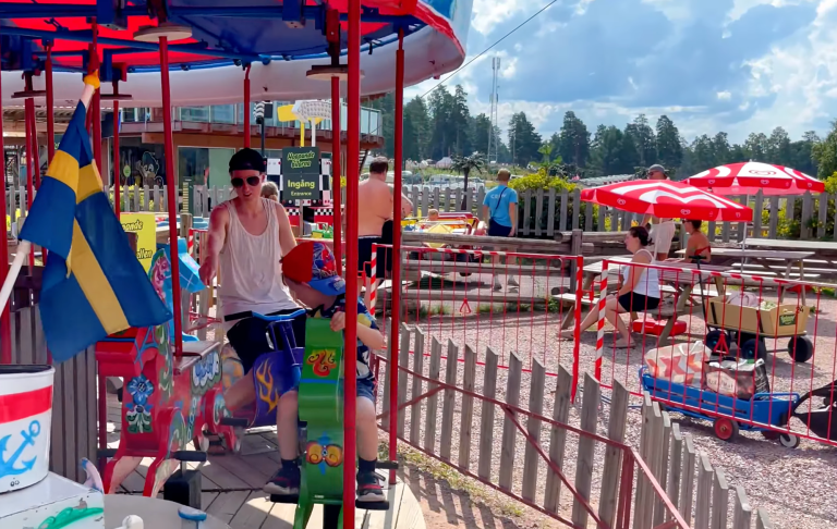 En ung man sitter på en karusell med svenska flaggan, omgiven av soliga parasoll och människor.