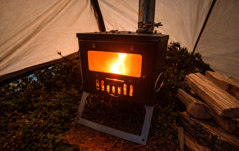 Hot tent kamin som brinner Vedspis med flammande eld inuti ett tältkåta, omgiven av ved och gröna växter.