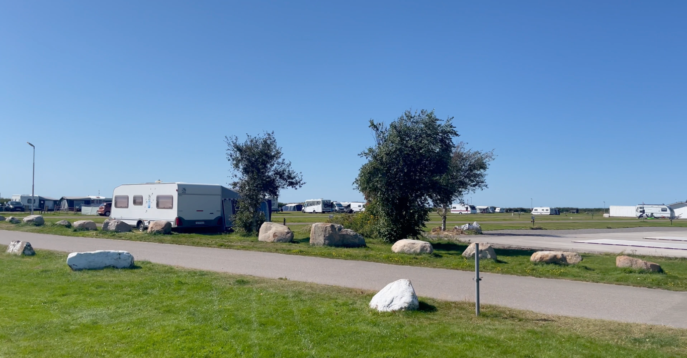 Björkängs Camping Campingområde med husvagnar, träd och stora stenar under klarblå himmel.