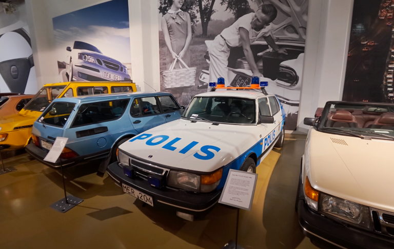Saab 900 Polisbil Vit polisbil med blå detaljer, omgiven av flera äldre bilar.