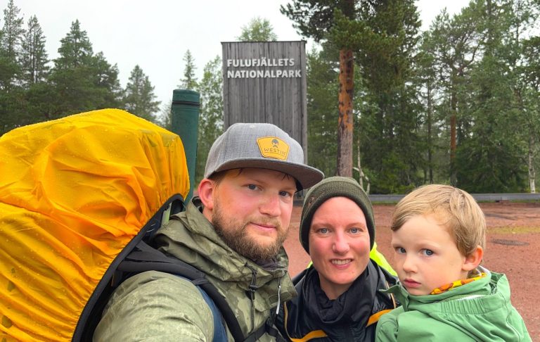Familj med ryggsäckar på vandring, står framför en stor informationstavla i skogen.