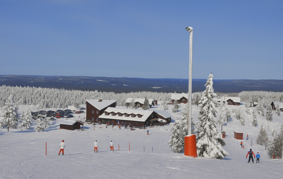 Snötäckt skidbacke med stugor och tallar i bakgrunden under klarblå himmel.