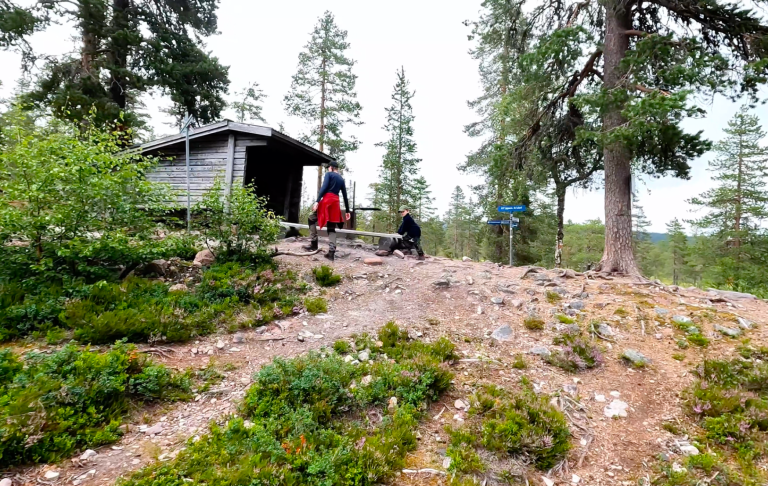 Två personer går mot en stuga omgiven av träd och grönska.