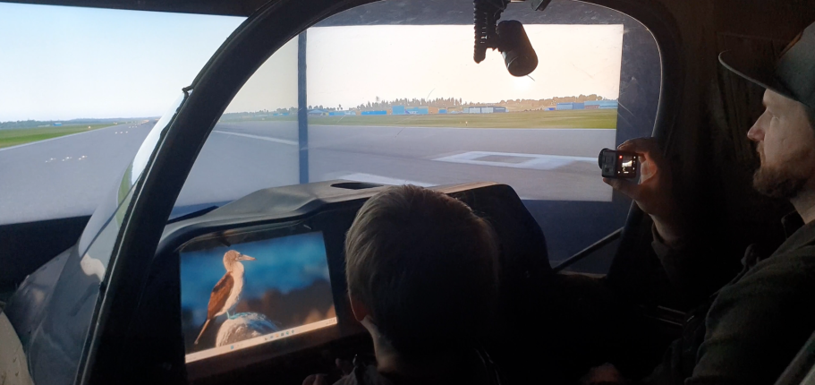 Flyger flygplan i flygsimulatorn på Aeroseum Två personer i en simulator med vy över en landsväg och en skärm som visar en fågel.