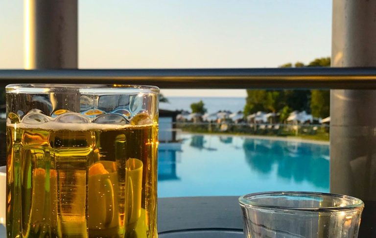 Ett glas öl framför en pool med havsutsikt vid solnedgång.