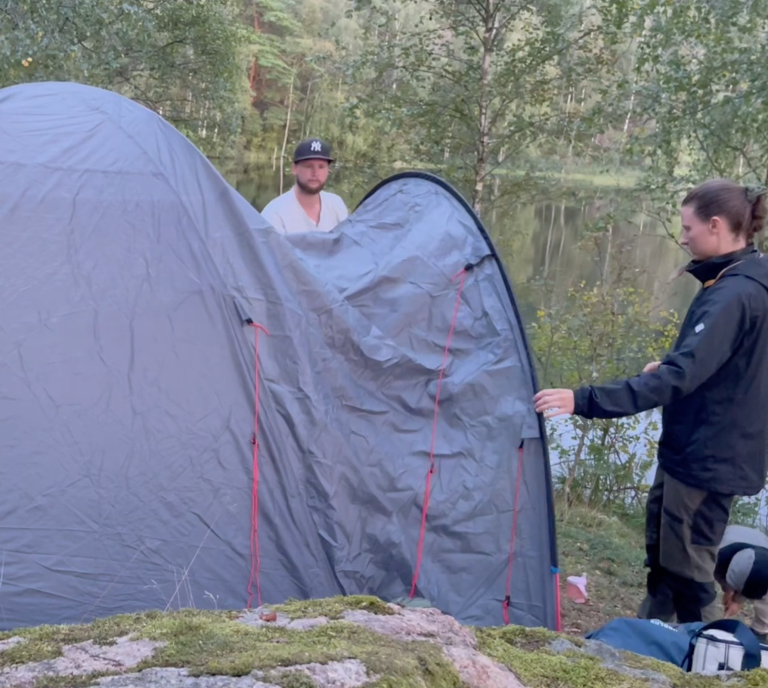 Två personer sätter upp ett tält vid en sjö i en naturskön miljö.
