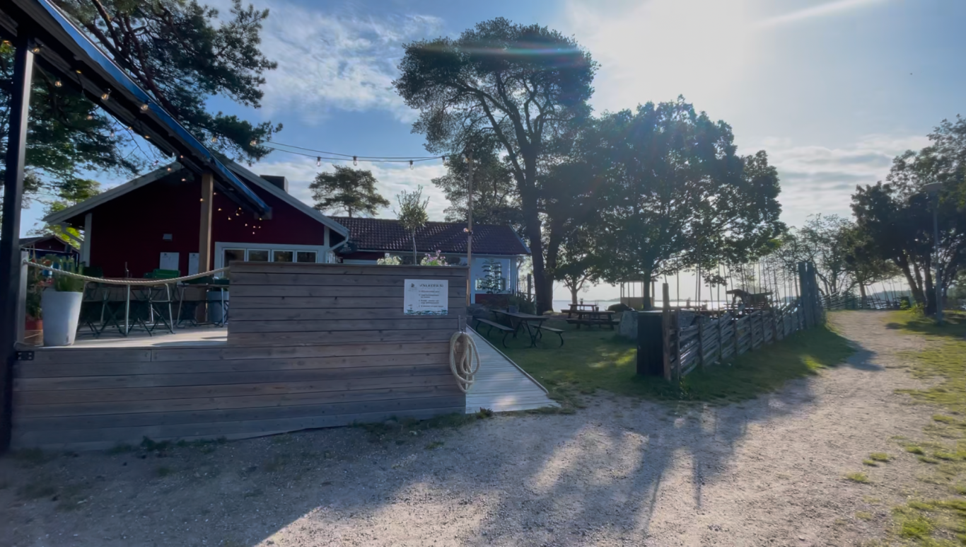 Algots på Udden Grusväg vid en stuga med träd och utsikt mot vattnet i bakgrunden.