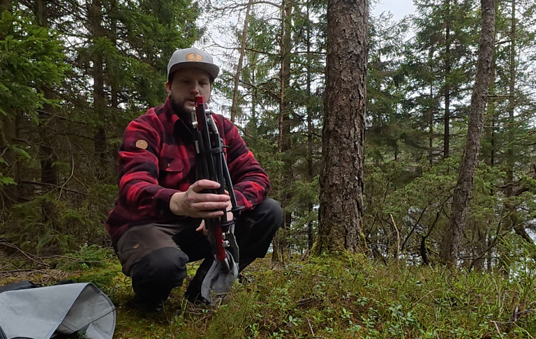 Man i rutig skjorta och keps sitter på knä i skogen, håller en ihopvikt ultralätt campingstol i handen.