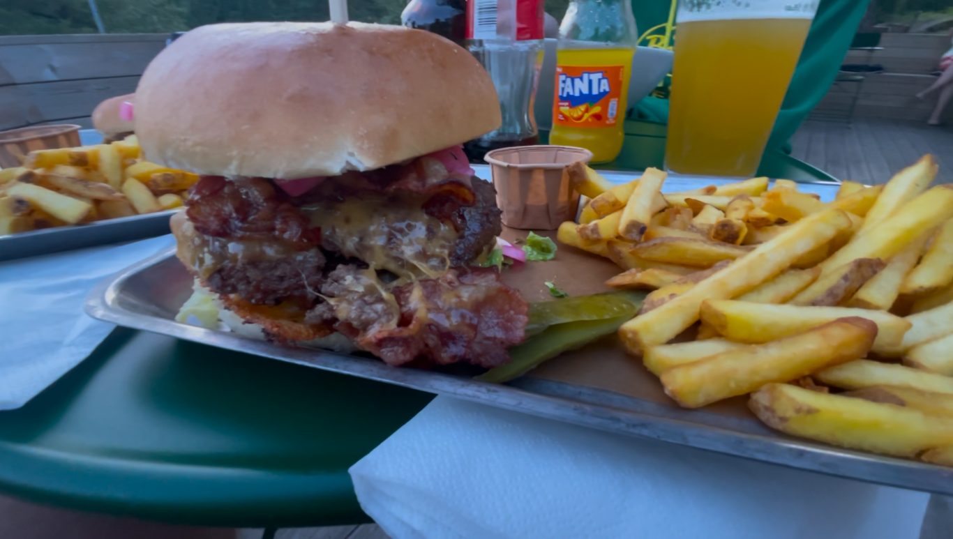 Maten på Algots på Udden En hamburgare med bacon, pommes frites och en läskig dryck på ett bord.