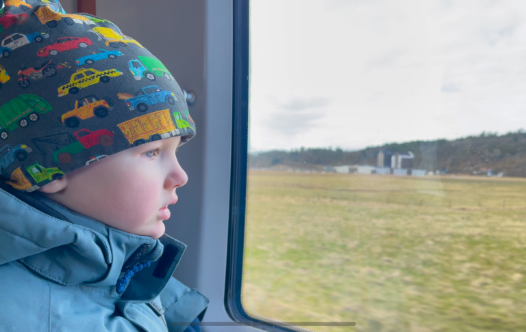 Litet barn tittar ut genom fönstret på en tågresa, landskap i bakgrunden.