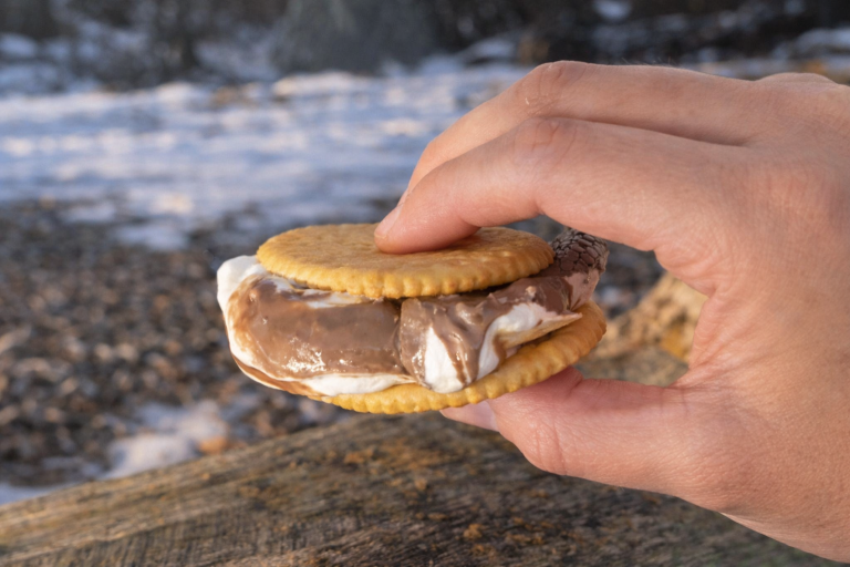 En hand håller en marshmallow, kex- och chokladfyllning mellan två kex.