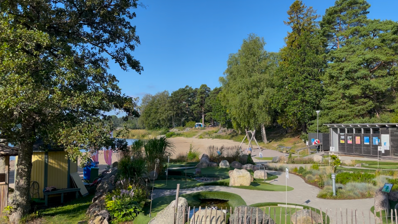 Minigolfbanan Lummig park med blomsterbäddar och träd, klarblå himmel i bakgrunden.