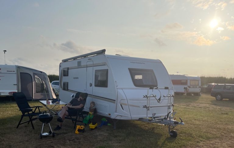 En vit husvagn står på en campingplats med en person som sitter utanför.