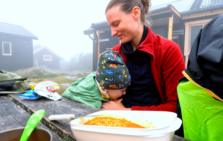 Kvinna sitter med ett barn, omgiven av natur, vid ett bord med mat.