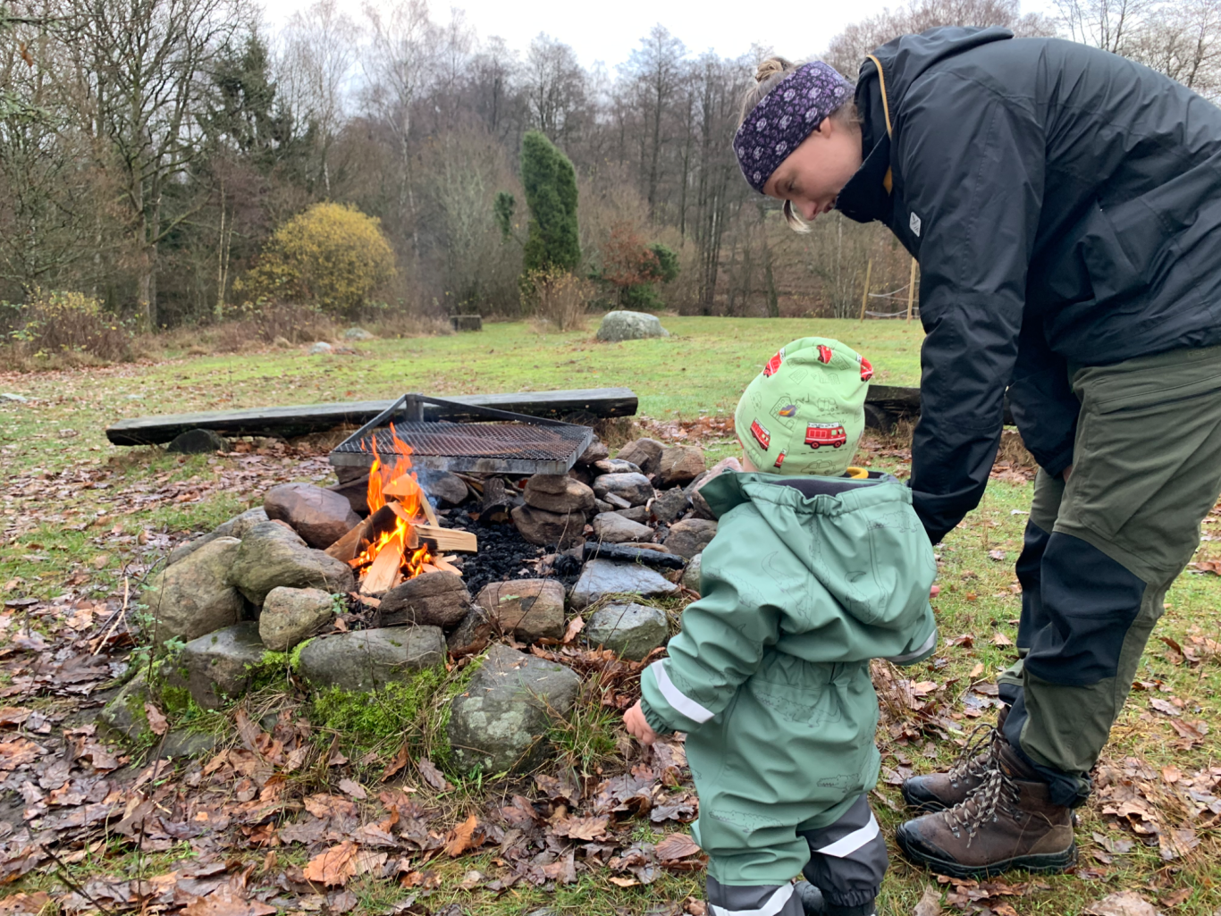 Grillar En vuxen och ett barn står vid en lägereld i naturen under en molnig dag.