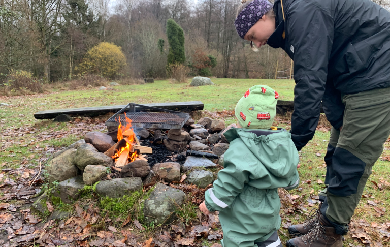 En vuxen och ett barn står vid en brasa i naturen, omgiven av träd och stenar.