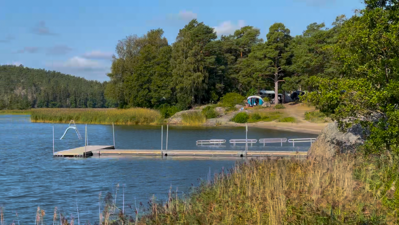 Badbryggan och stranden Brygga vid en sjö omgiven av träd och en campingplats i bakgrunden.