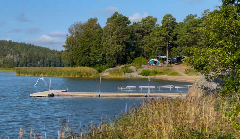 Sandstranden på Trosa Havsbad och Camping Brygga vid en sjö omgiven av träd och gräs, med campingplats i bakgrunden.