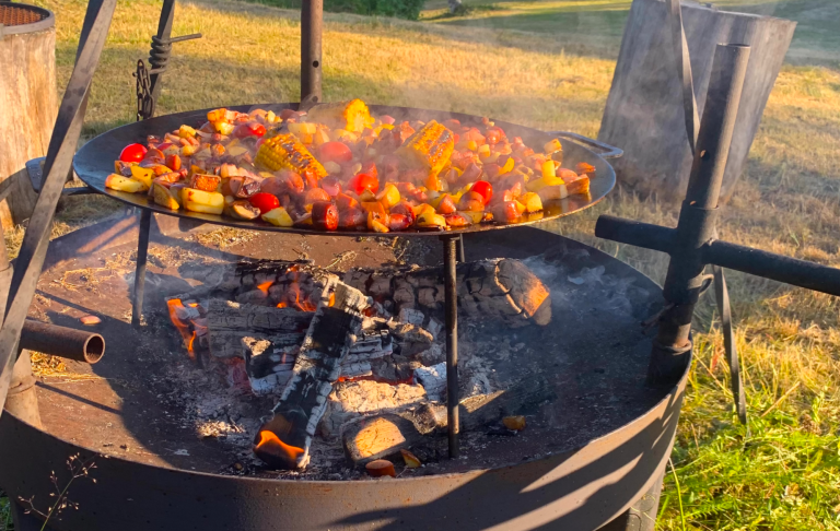 Grill med grönsaker och kött som tillagas över öppen eld på en utomhusplats.