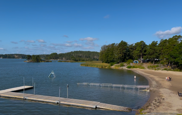 Lugnt vattendrag med bryggor, sandstrand och grönska under en klarblå himmel.