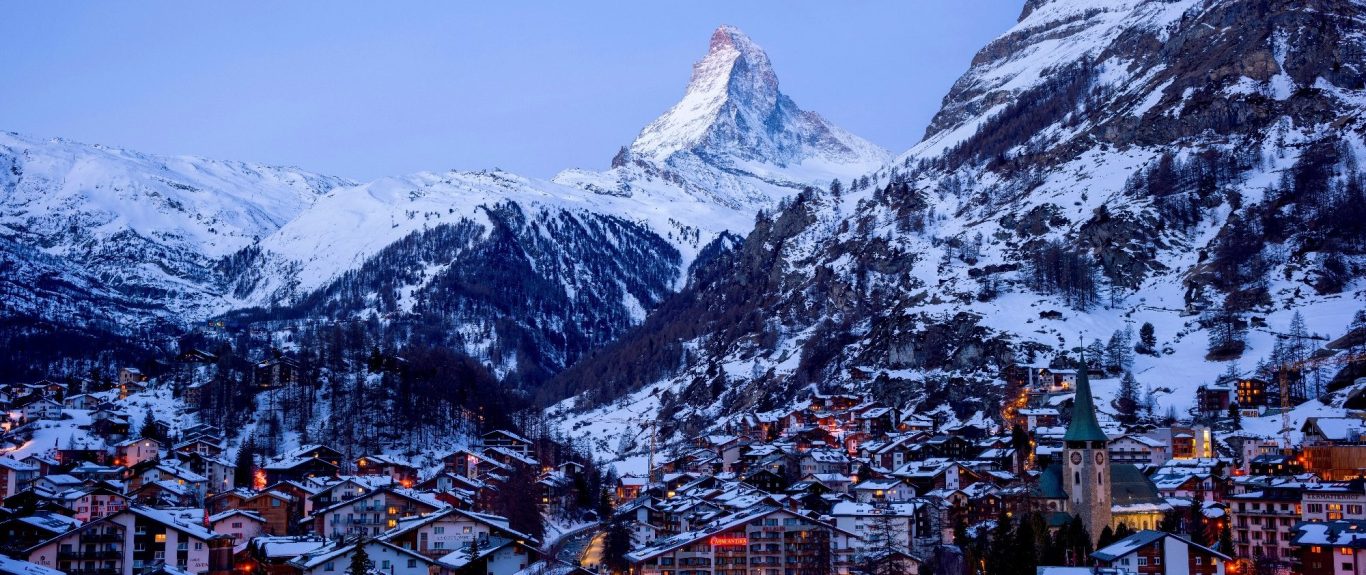 Snötäckta alpberg med Matterhorn och en by med ljus under skymningen.