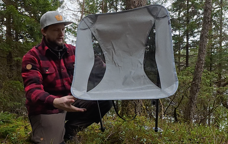 En man i röd och svart rutig skjorta håller upp en campingstol i skogen.