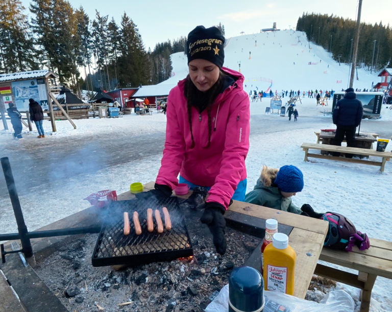 Kvinna grillar korv på en utekamp, vinterlandskap och snötäckta backar i bakgrunden.