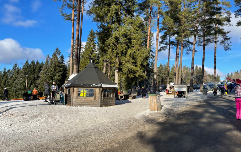 Kiosk vid skidområde omgiven av höga träd och snö, med besökare i bakgrunden.