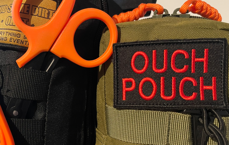 Grön väska med texten "OUCH POUCH" och en orange sax bredvid.