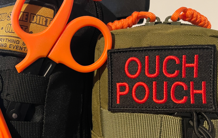 Grön väska med texten "OUCH POUCH" och orange saxar synliga.