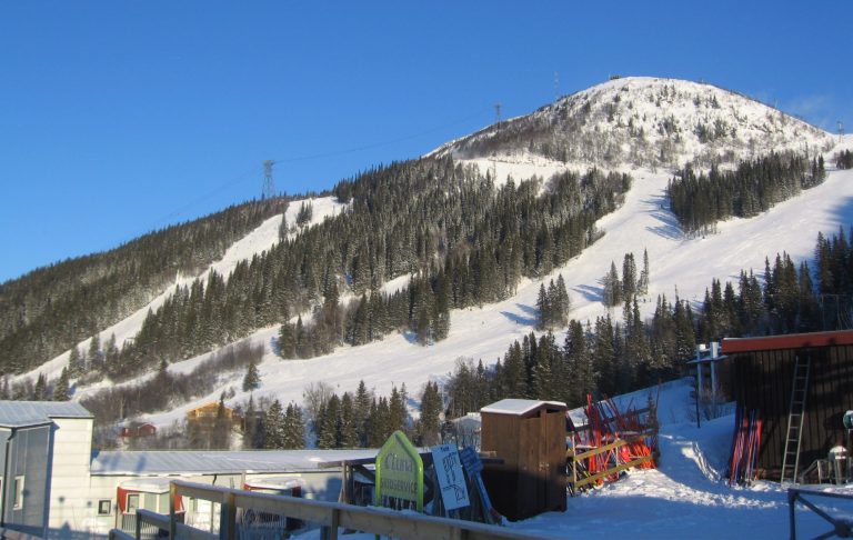 Snötäckt skidbacke med träd, byggnader och klarblå himmel.