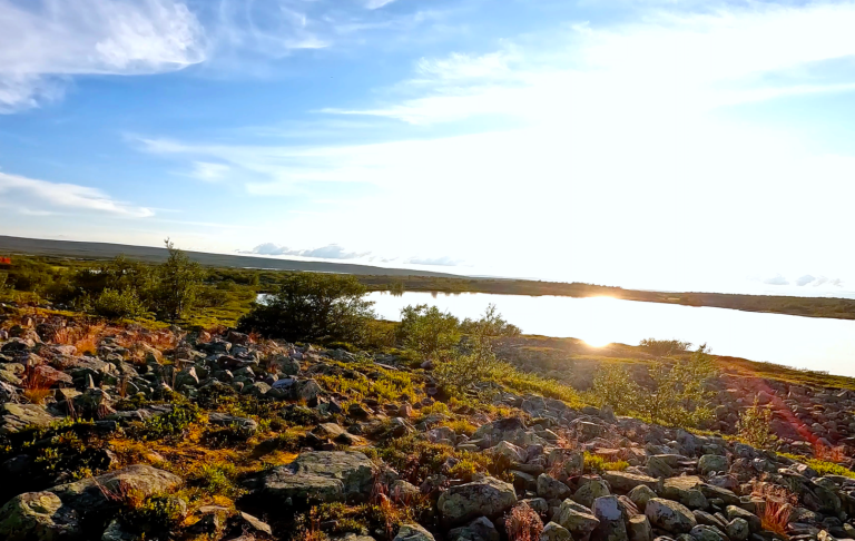 Solnedgång över en sjö med klippor och vegetation i förgrunden.