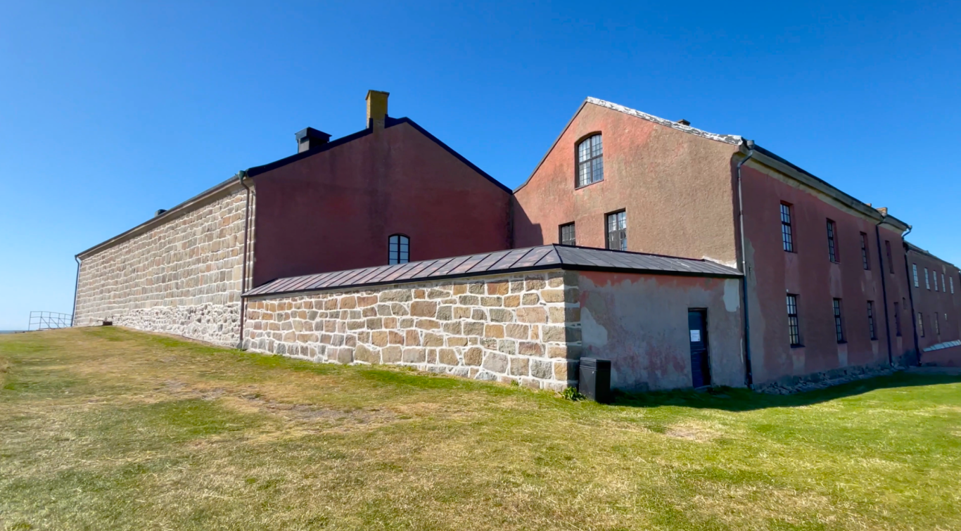 Varbergs Fästning Gamla byggnader med stenar och puts, belägna på en grön sluttning under klar himmel.