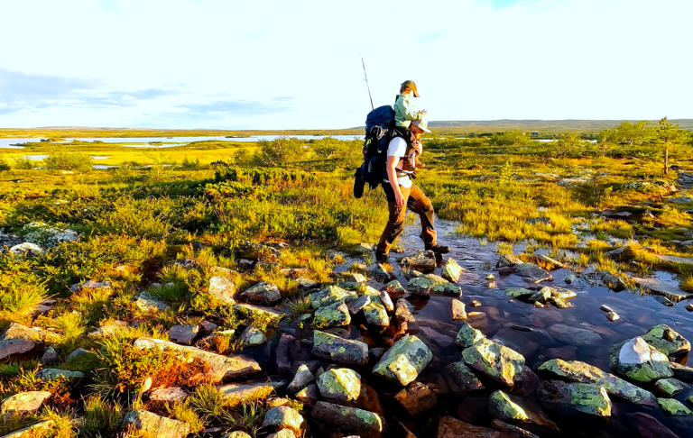 Person vandrar med ryggsäck över stenig terräng i en grön landskap.