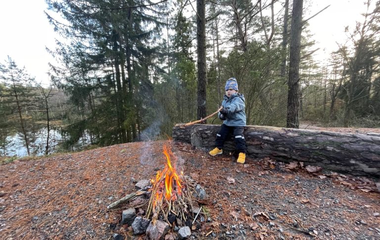 Ett barn sitter vid en brasa i skogen, med en pinne i handen.