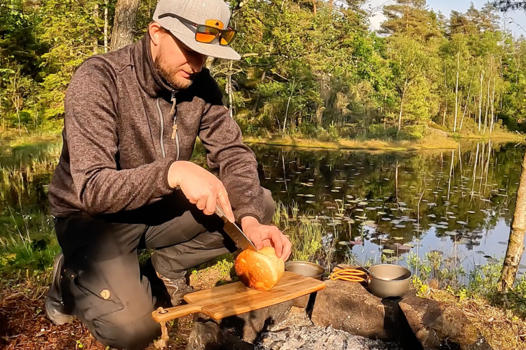 En person skär bröd vid en lägereld nära en spegelblanka sjö.