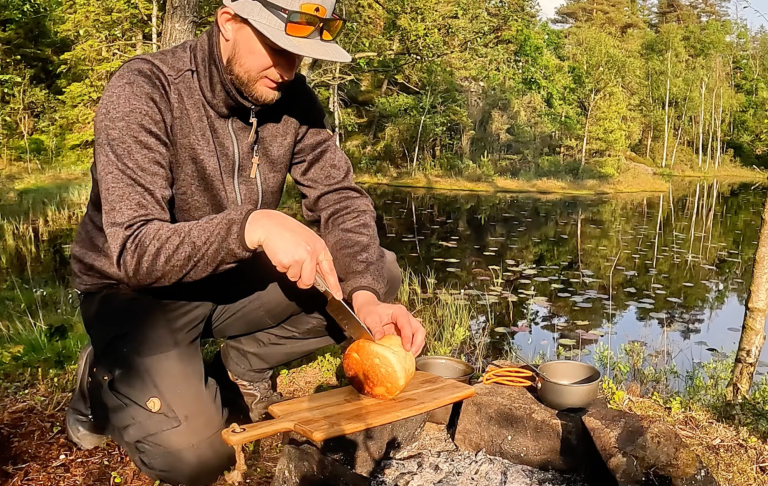 En person skär ett bröd vid en sjö, omgiven av grönska och natur.