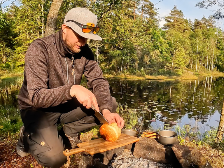 En person skär ett bröd vid en sjö, omgiven av skog och soligt väder.