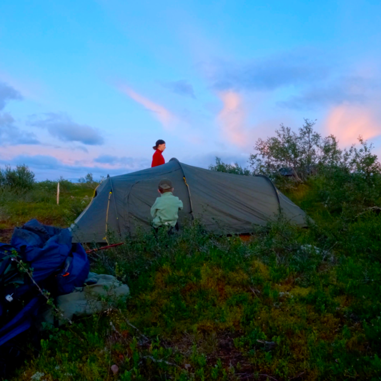 Två personer sätter upp ett tält i en naturskön omgivning under blå himmel.
