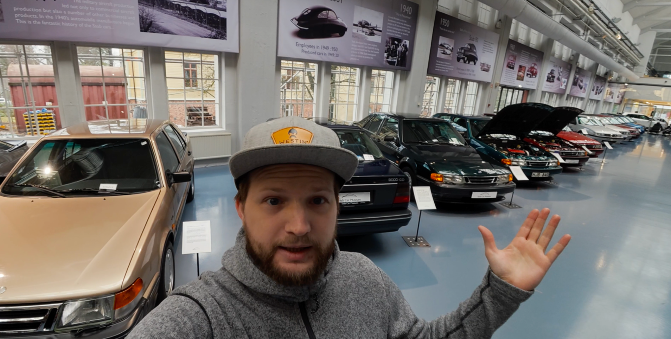 Johannes Framför Saab 9000 En person står framför en utställning av flera bilar inomhus.
