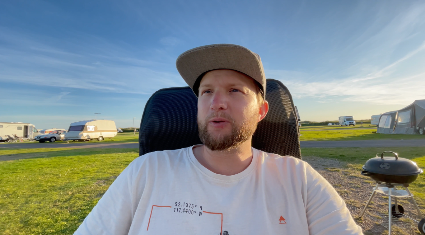 Johannes En man sitter på en stol utomhus, med grönska och fordon i bakgrunden.