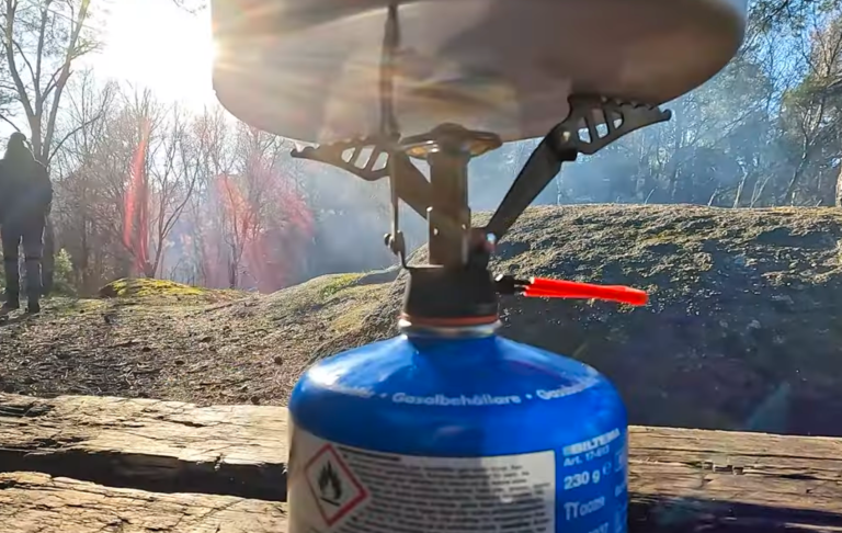 Campingkök med gasflaska, i bakgrunden en suddig person och solens strålar.