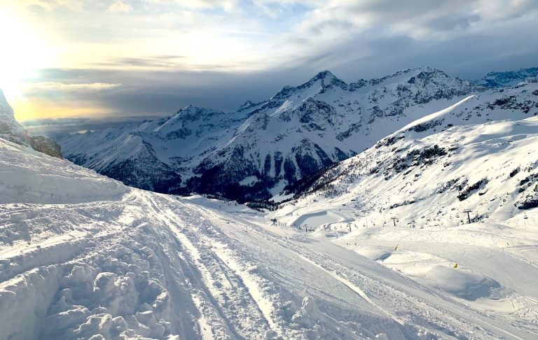 Vinterlandskap med snötäckta alpberg och skidspår under en ljus himmel.