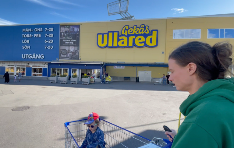 Kvinna står bredvid en barnvagn utanför Ullareds butik. Klara och soliga förhållanden.