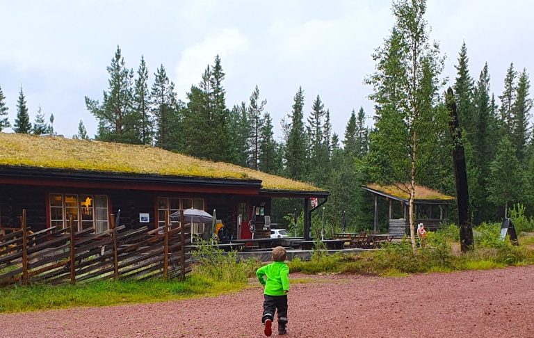 Vandrare med grön tröja vid stugor omgivna av tallar och grönskande natur.