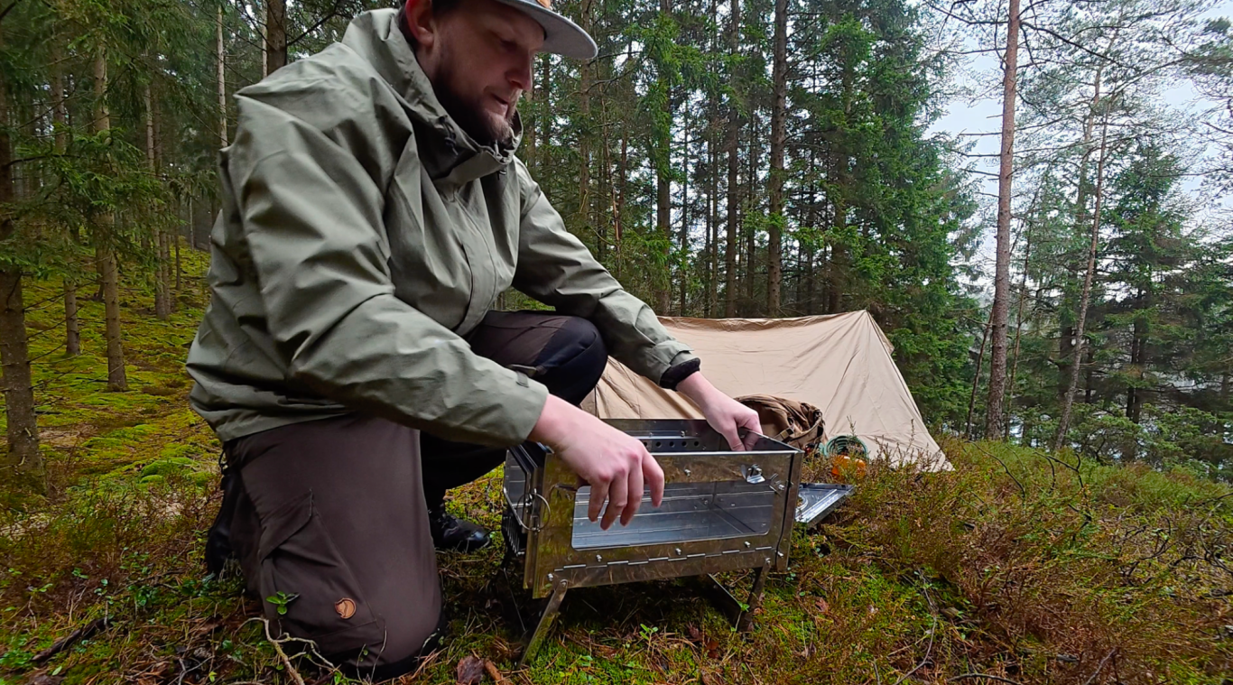 Monterar kaminen En person i skogsmiljö förbereder en ugn framför ett tält.
