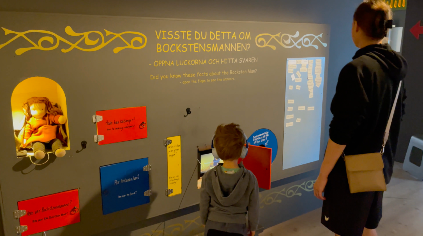 Bockstensmannen för barn En vuxen och ett barn står framför en interaktiv utställning med färgglada skärmar.