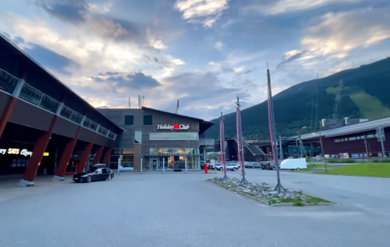 Ett modernt komplex med butiker och parkering, omgiven av berg och himmel med moln.